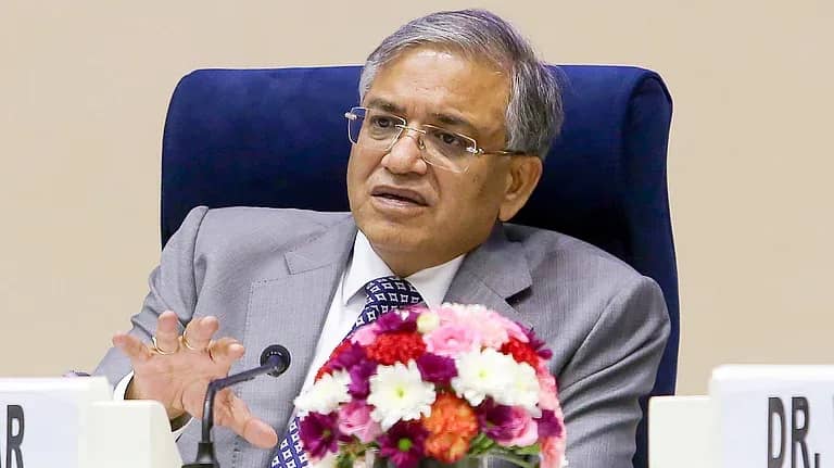 ایک شخص سوٹ اور ٹائی پہنے، میز پر بیٹھا، مائیکروفون کے قریب ہاتھ ہلاتے ہوئے