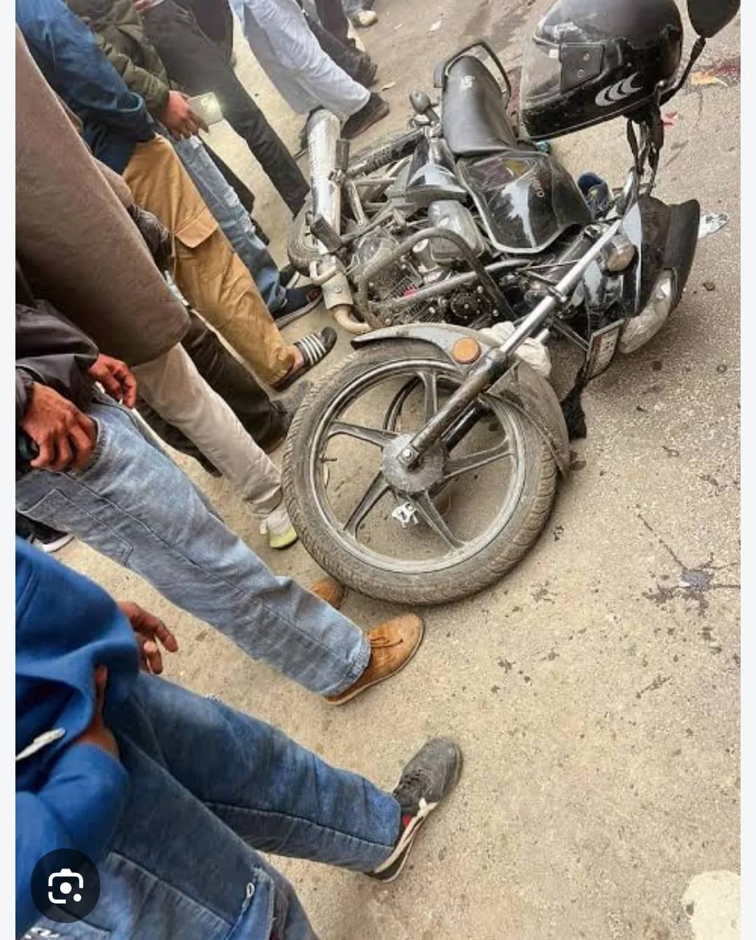ایک موٹر سائیکل گرا ہوا ہے اور کچھ لوگ اس کے ارد گرد کھڑے ہیں