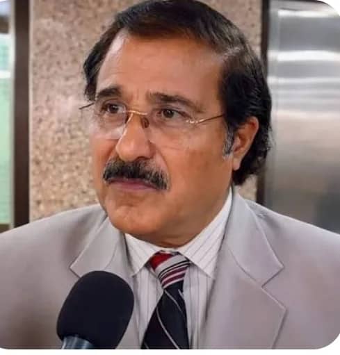 ایک شخص مائیکروفون کے سامنے کھڑا ہے، جس نے سوٹ اور ٹائی پہنی ہوئی ہے۔
