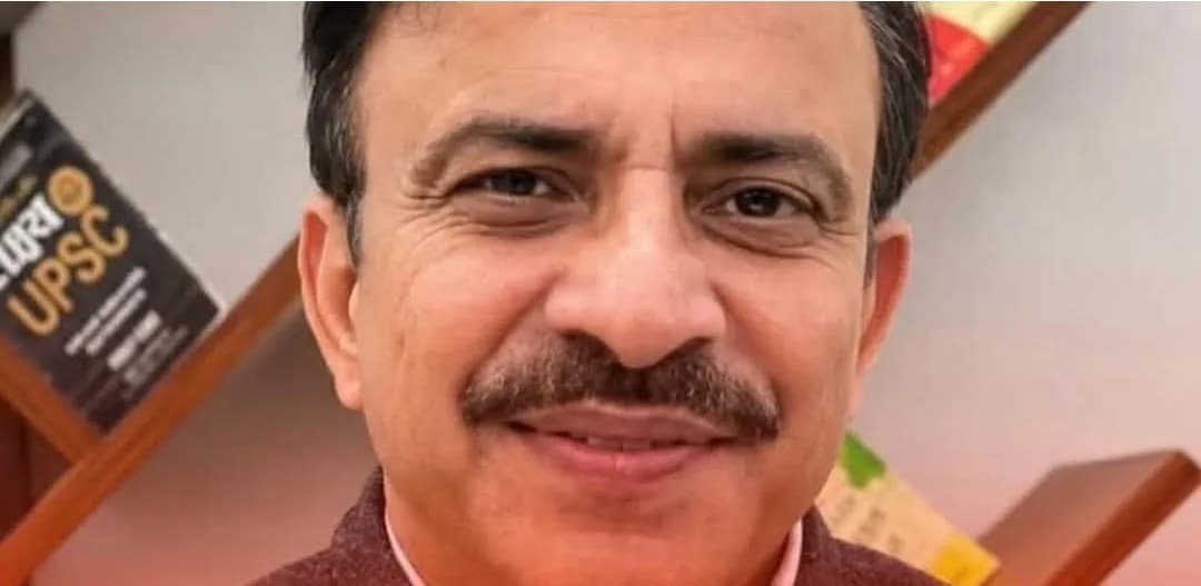 ایک درمیانی عمر کا مرد کیمرے کی طرف مسکرا رہا ہے، اس کے پیچھے کتابوں کی الماری ہے۔