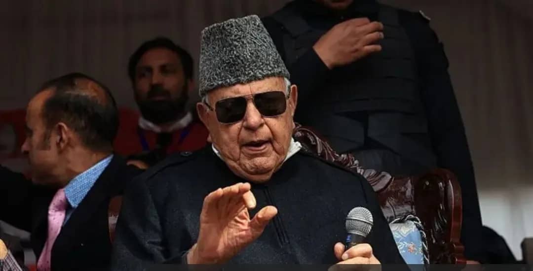 ایک بزرگ شخص کالی ٹوپی اور دھوپ کے چشمے پہنے مائیکرو فون پر بات کر رہا ہے۔