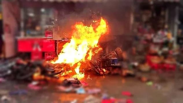 ایک سڑک پر آگ لگی ہوئی ہے، جس کے پس منظر میں سرخ رنگ کے کریٹ اور دھندلی عمارتیں ہیں۔