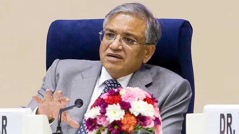 ایک شخص سوٹ اور ٹائی پہنے، میز پر بیٹھا، مائیکروفون کے قریب ہاتھ ہلاتے ہوئے