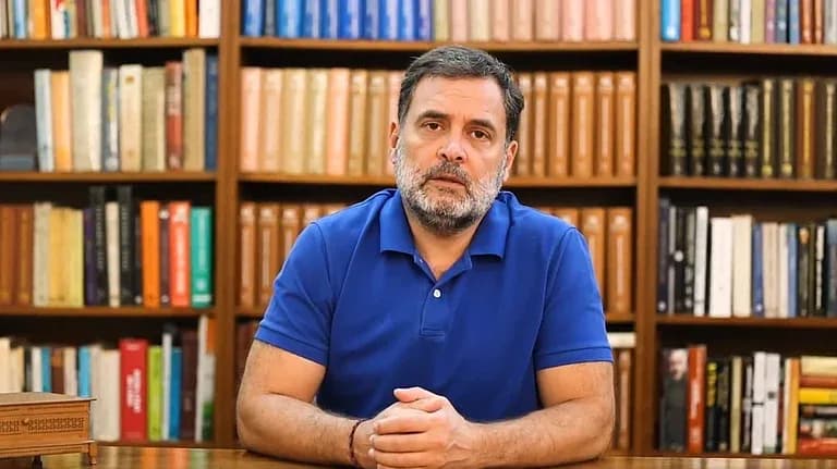 ایک شخص نیلے رنگ کی پولو شرٹ پہنے، داڑھی کے ساتھ، کتابوں کی الماری کے سامنے بیٹھا ہے۔