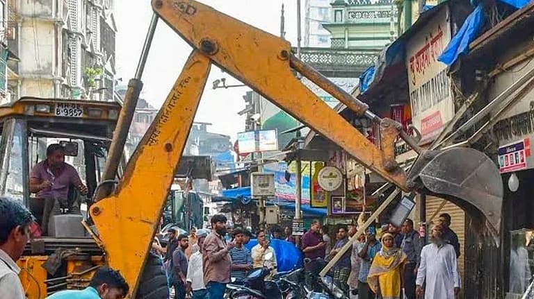 ایک پیلے رنگ کا بلڈوزر دکانوں کو گرا رہا ہے جبکہ لوگ دیکھ رہے ہیں۔